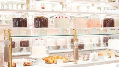 A bakery display with cakes under glass domes, assorted pastries, and cookies on white stands evokes the charm of the Best Disney Resorts for Evening Activities, set against white shelves filled with jars and dishes in a bright, elegant setting.