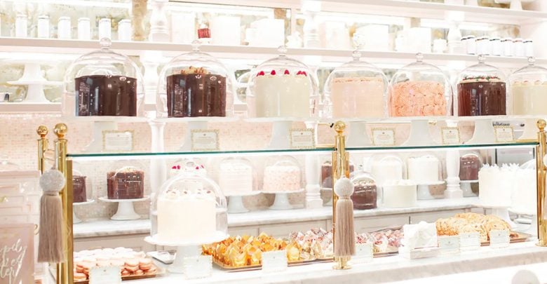 A bakery display with cakes under glass domes, assorted pastries, and cookies on white stands evokes the charm of the Best Disney Resorts for Evening Activities, set against white shelves filled with jars and dishes in a bright, elegant setting.