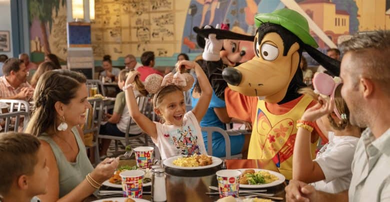 At Walt Disney World, a family sits at a restaurant table, smiling and enjoying a meal. A person dressed as a cartoon dog character playfully interacts with a young girl wearing mouse ear headbands. Colorful cups dot the table, adding to the lively and cheerful atmosphere.