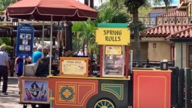A colorful food cart boasting "Spring Rolls" and Magic Kingdom Snacks sits outdoors, adorned with a decorative umbrella. In the background, buildings and palm trees add to the charm as a few people stroll by.