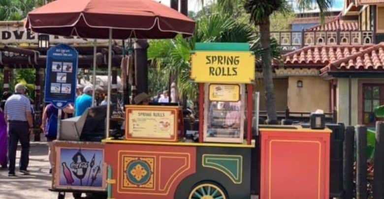 A colorful food cart boasting "Spring Rolls" and Magic Kingdom Snacks sits outdoors, adorned with a decorative umbrella. In the background, buildings and palm trees add to the charm as a few people stroll by.