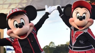 Mickey Mouse and Minnie Mouse, dressed in matching red and black athletic outfits, hold hands and raise their arms in celebration outdoors during the Disney Princess Half Marathon Weekend.