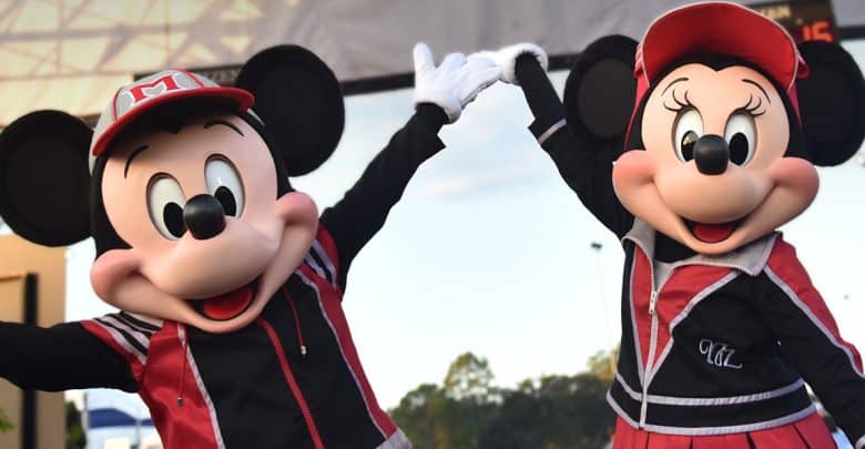 Mickey Mouse and Minnie Mouse, dressed in matching red and black athletic outfits, hold hands and raise their arms in celebration outdoors during the Disney Princess Half Marathon Weekend.
