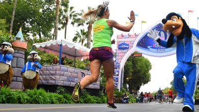 A runner in athletic gear gives a high-five to someone in a Goofy costume near the finish line, with Chip and Dale mascots cheering in the background during the Disney Princess Half Marathon Weekend.