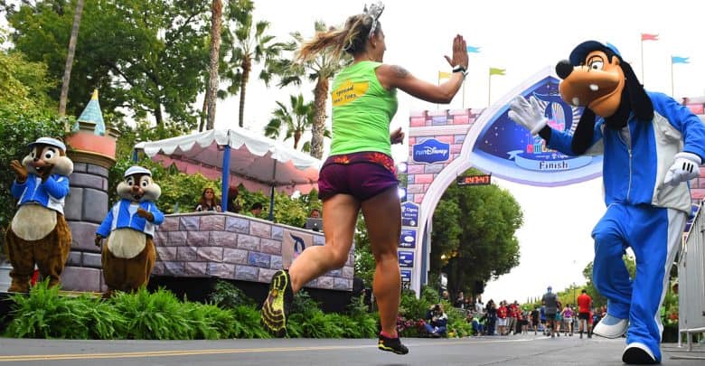 A runner in athletic gear gives a high-five to someone in a Goofy costume near the finish line, with Chip and Dale mascots cheering in the background during the Disney Princess Half Marathon Weekend.