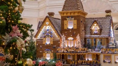 A detailed gingerbread house stands near a decorated Christmas tree at the Grand Floridian Resort & Spa. The house, with its snowy rooftops, lit windows, and festive decorations, creates a cozy holiday scene indoors. A balcony and miniature figures add extra charm.