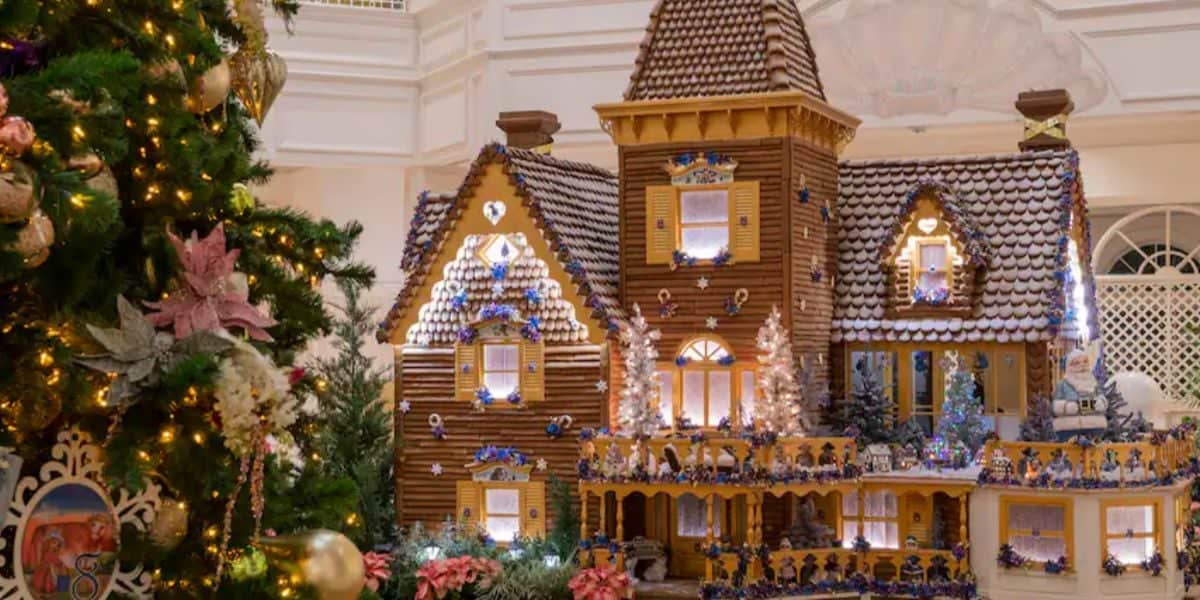 A detailed gingerbread house stands near a decorated Christmas tree at the Grand Floridian Resort & Spa. The house, with its snowy rooftops, lit windows, and festive decorations, creates a cozy holiday scene indoors. A balcony and miniature figures add extra charm.