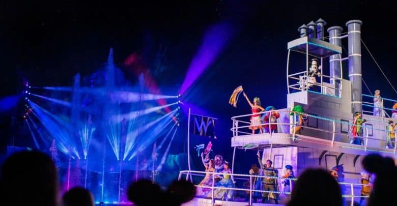 A lively nighttime performance at Disney's Hollywood Studios features colorful lights and water fountains in the background. Performers dressed in vibrant costumes are on a large, white boat decorated with lights. The scene is energetic, filled with color and motion.