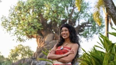 A person dressed in a Polynesian-inspired costume reminiscent of Disney Princesses stands smiling in front of a large artificial tree at an amusement park, surrounded by lush greenery and palm trees. The sky is clear, indicating a sunny day.