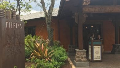 At Walt Disney World, the entrance to Tiffins and Nomad Lounge features a large sign and decorative metal sculpture. Two people stand at the reservation podium near the doorway, surrounded by lush greenery, capturing the essence of a dream Disney vacation.