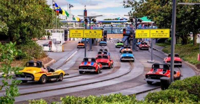 Numerous colorful, small cars navigate a track at an amusement park. The track is surrounded by greenery and various flags, with a gated entrance visible in the background. As one of the important rides in Magic Kingdom, the atmosphere is lively, with people enjoying their joyful rides.