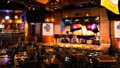 The image captures a dimly lit, rustic bar at Walt Disney World, featuring wooden tables and chairs facing a stage with two grand pianos. Neon signs glow on the walls, and banners hang from the ceiling, creating an inviting atmosphere perfect for adults seeking a lively evening.