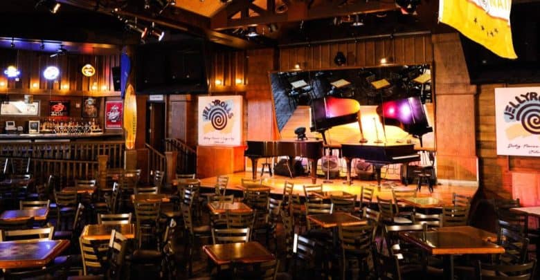 The image captures a dimly lit, rustic bar at Walt Disney World, featuring wooden tables and chairs facing a stage with two grand pianos. Neon signs glow on the walls, and banners hang from the ceiling, creating an inviting atmosphere perfect for adults seeking a lively evening.