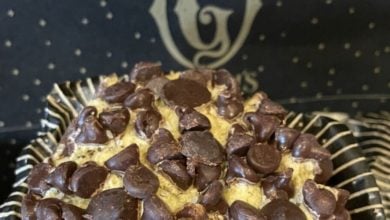 A close-up of a muffin topped with numerous chocolate chips brings to mind the delightful world of Disney Snacks. The muffin is in a dark-colored wrapper with a striped pattern, while a blurred background reveals part of a logo or design in a contrasting color.