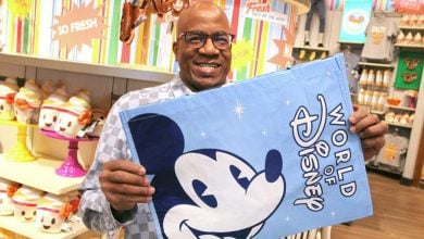 A man smiles while holding a blue "World of Disney" bag featuring Mickey Mouse, enjoying the colorful displays and Disney merchandise—proof that staying cool at Disney is all about finding magic indoors.