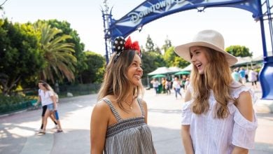 Two young women smile and chat under a “Downtown Disney” sign—one in Minnie Mouse ears and a sundress, the other in a white hat and off-shoulder top—capturing the fun vibe often compared in Disney Springs vs Downtown Disney debates.
