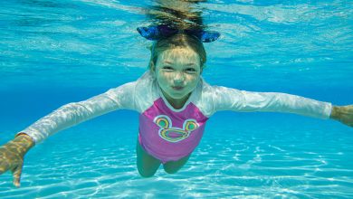 A young girl in a pink swimsuit with a cartoon face swims underwater in a clear blue pool, arms outstretched and hair floating, with sunlight reflecting on the pool floor—perfect for staying cool at Disney.