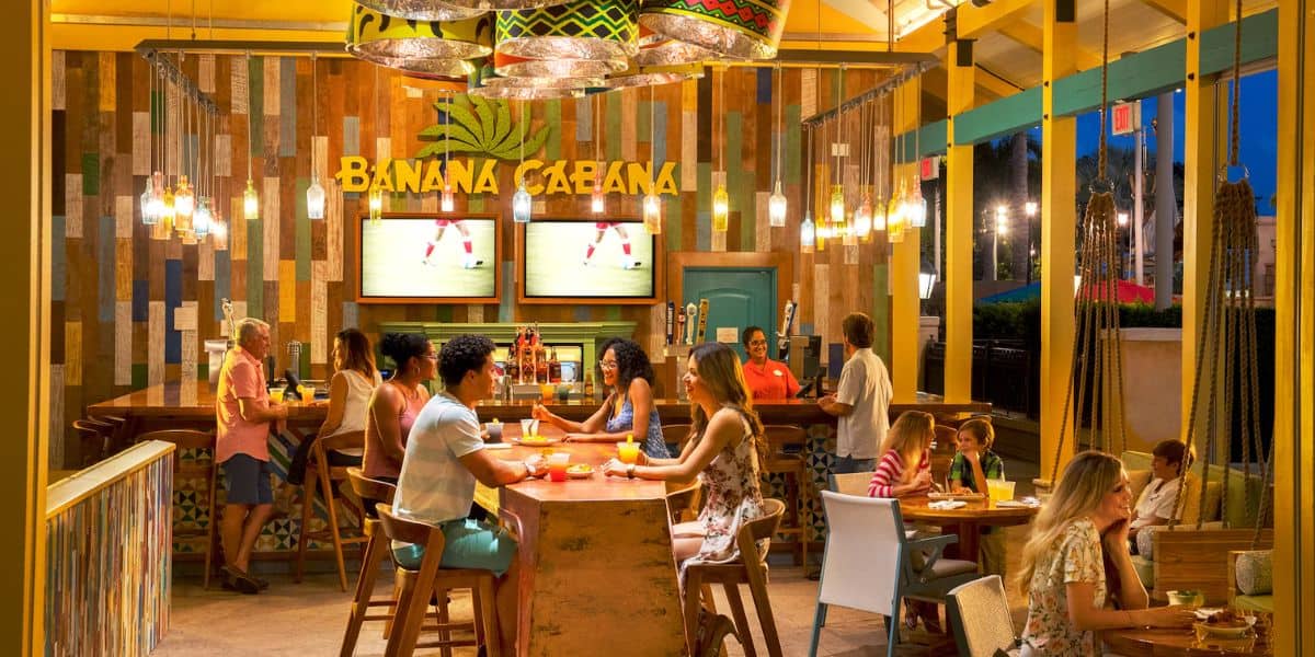 Diners enjoy the tropical-themed Banana Cabana, nestled among Disney Resort Lounges. The vibrant space boasts colorful wooden decor, ceiling lights, and two TVs. A bar and diverse seating areas enhance the relaxed, lively atmosphere, perfect for a magical escape.