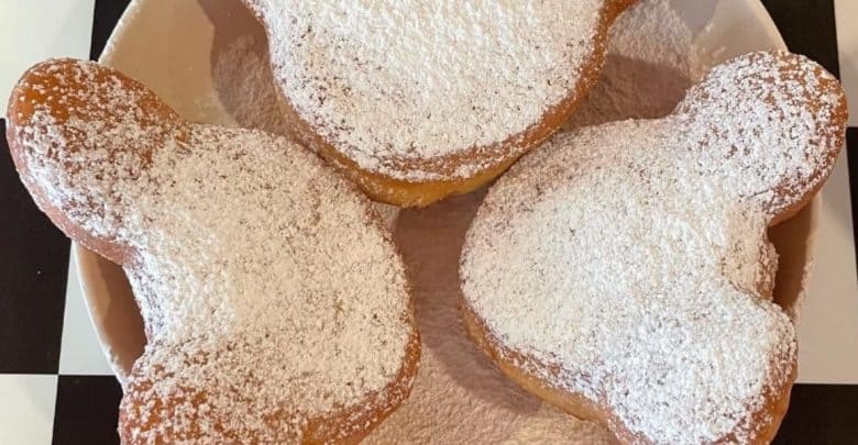 Three Mickey Mouse-shaped beignets, a delightful example of cheap Disney snacks, are covered in powdered sugar and served on a plate with a black and white checkered background.