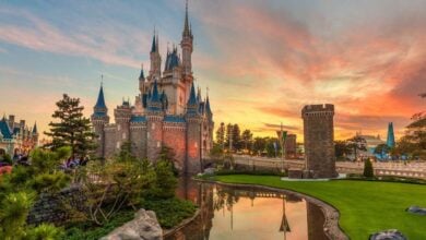 A fairytale castle with blue spires stands beside a moat, surrounded by trees and green lawns, illuminated by a colorful sunset sky. People gather near the castle, enjoying the scenic view.