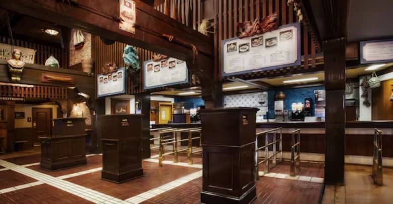 The vintage fast-food restaurant, reminiscent of days gone by, boasts wooden decor and illuminated menu boards above the counter area. Downstairs, an organized path with metal rails guides patrons forward, while upstairs dining options at Walt Disney World elevate the experience further.