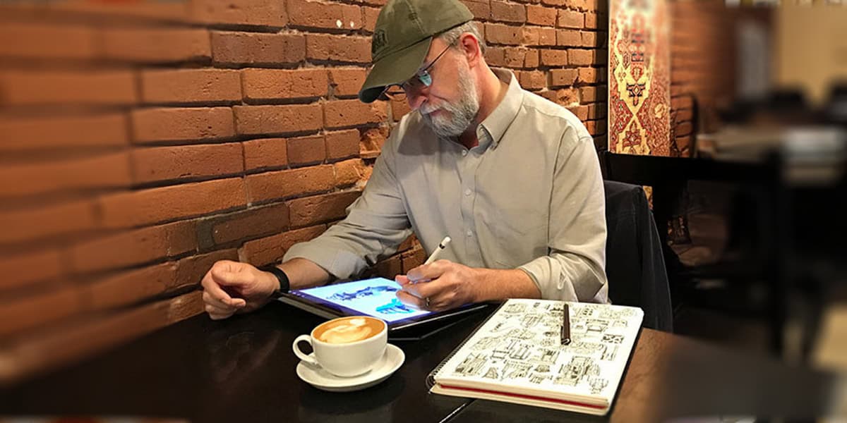 A bearded man in a cap sketches on a tablet at a café, coffee and notebook beside him, dreaming to become a Disney Imagineer. A brick wall and warm decor set the creative scene.