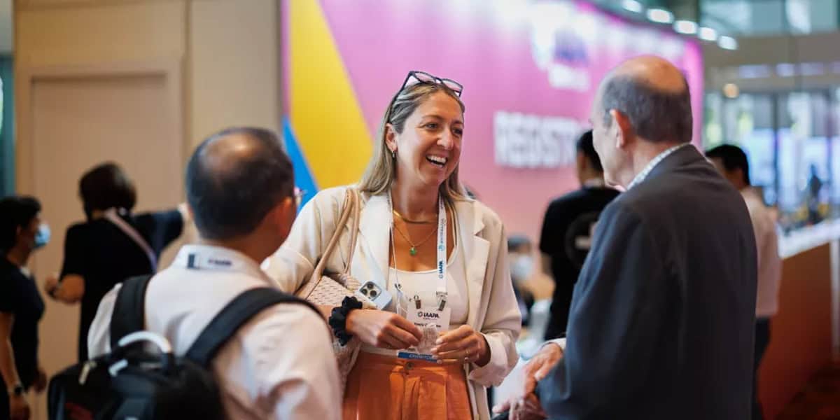 A woman smiles and chats with two men at a conference, standing indoors near a colorful registration sign. Other attendees are visible, discussing how to become a Disney Imagineer in the vibrant, engaging atmosphere.