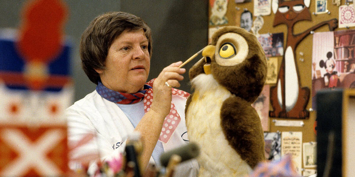 A person in a white coat and scarf works on a brown and white owl puppet in a vibrant Imagineering workshop