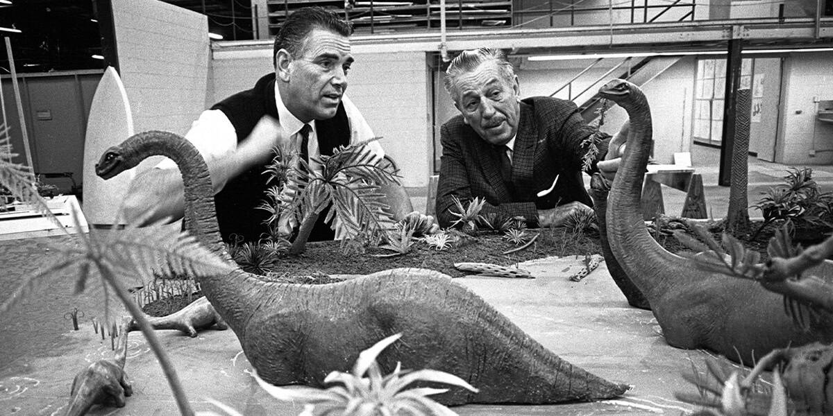 Walt Disney examines a detailed miniature diorama featuring model dinosaurs and plants.