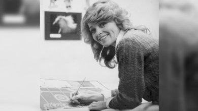 A woman with wavy hair smiles at the camera while drawing on a large piece of paper.