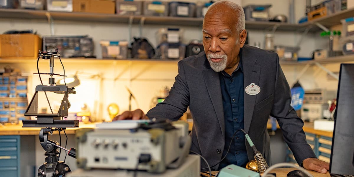 Lanny Smoot in a suit works in an electronics lab, surrounded by equipment and tools.