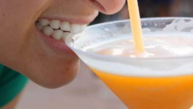 A close-up of a person smiling and drinking an orange beverage from a glass using a straw. The person's teeth and part of their face are visible.