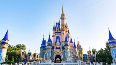 Cinderella Castle at Walt Disney World, featuring tall spires with blue and gold accents, stands under a clear blue sky, surrounded by decorative banners and greenery in this location where disney world open carry is not permitted.