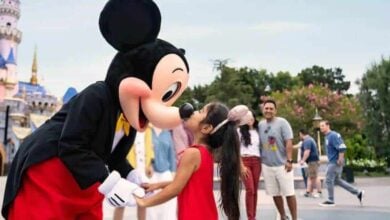 A young girl in a red dress smiles as she meets Mickey Mouse at Disney World, with other guests and Cinderella Castle behind them.