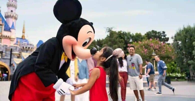 A young girl in a red dress smiles as she meets Mickey Mouse at Disney World, with other guests and Cinderella Castle behind them.