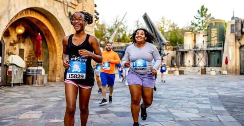 People are happily running in a themed park area, wearing race bibs with numbers. Two women in the foreground are smiling and jogging side by side on a paved pathway, their footsteps echoing amidst futuristic buildings reminiscent of Disney's sci-fi fantasies.