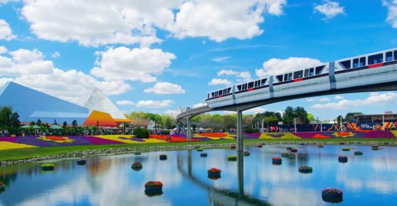 The Disney monorail glides over EPCOT’s pond, surrounded by floating flower beds and vibrant pavilions beneath a bright sky.