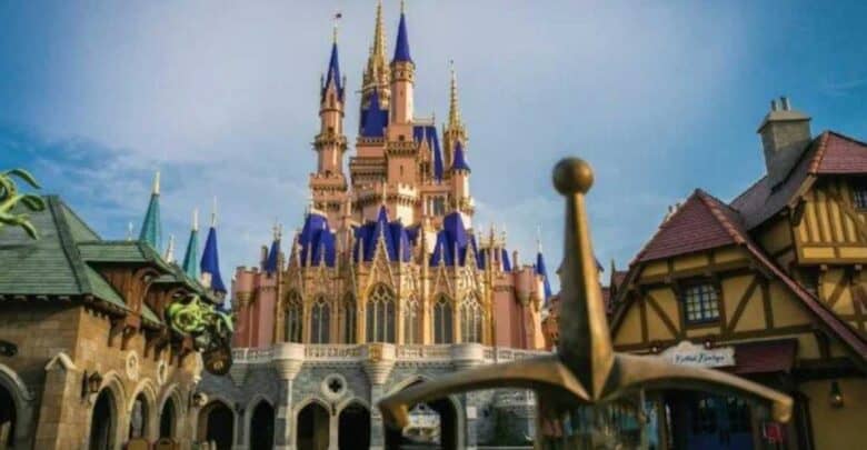A view of a fairytale castle with pink walls and blue spires, surrounded by fantasy-themed buildings, with a sword in a stone in the foreground under a blue sky.