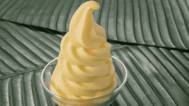 A swirl of creamy yellow soft-serve ice cream, reminiscent of iconic Disney snacks, sits in a clear glass cup. The background features a textured pattern resembling large, overlapping green leaves.