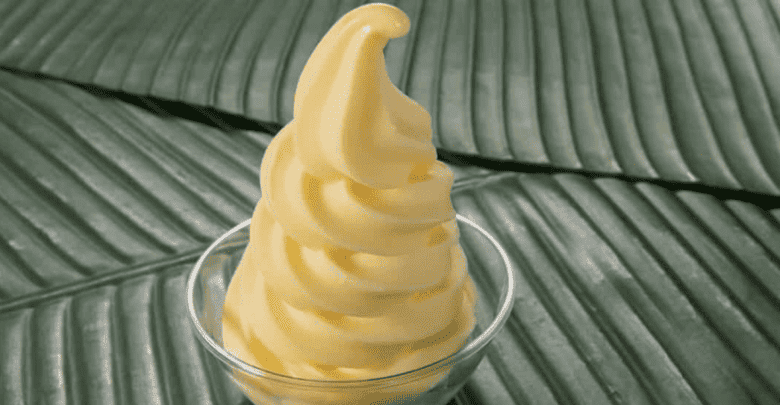 A swirl of creamy yellow soft-serve ice cream, reminiscent of iconic Disney snacks, sits in a clear glass cup. The background features a textured pattern resembling large, overlapping green leaves.