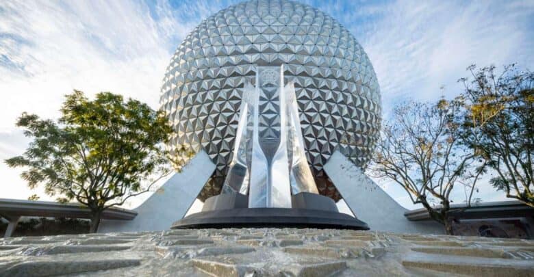 Low-angle view of a large geodesic sphere with a triangular pattern, surrounded by trees under a blue sky with wispy clouds. The foreground features a textured walkway leading up to the sphere.