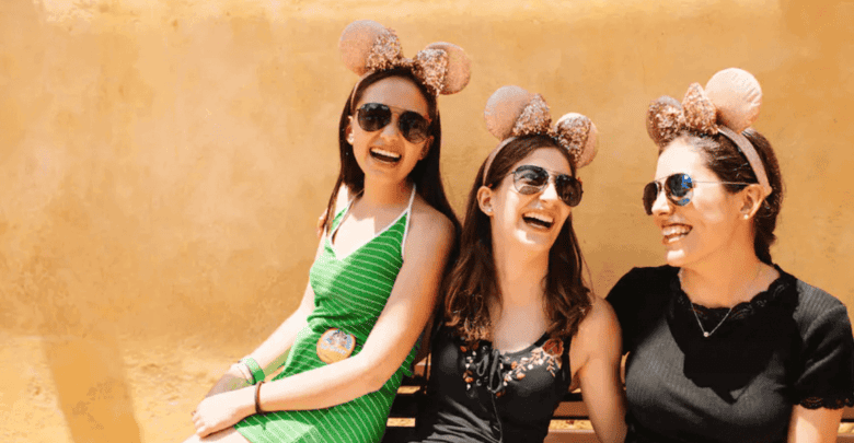 Three women wearing Mickey Mouse ear headbands and sunglasses are sitting on a bench against a warm-toned background, smiling and enjoying each other's company on a sunny day. Their laughter is contagious, making it clear they are having the time of their lives with some Walt Disney World tips in mind.