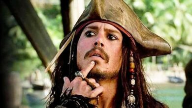 A person in a pirate costume with a tricorn hat touches their chin thoughtfully, wearing beads in their long hair and various rings, with a tropical background behind them.