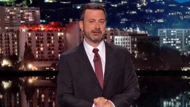 A man in a dark suit and red tie stands on a stage with a cityscape backdrop at night, illuminated buildings visible behind him. He appears to be hosting or giving a speech, with his hands clasped in front.