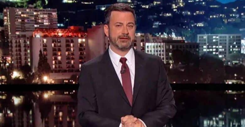 A man in a dark suit and red tie stands on a stage with a cityscape backdrop at night, illuminated buildings visible behind him. He appears to be hosting or giving a speech, with his hands clasped in front.