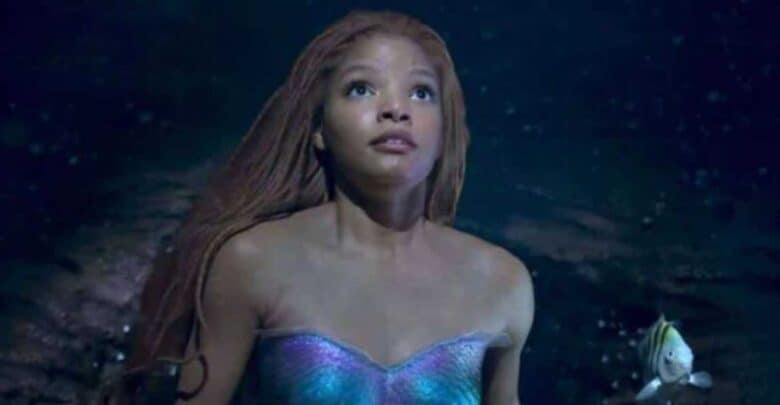 A young woman with long reddish hair and a shimmering mermaid tail looks upward underwater, with a small colorful fish swimming beside her. The scene is dark, illuminated by soft, blue light.