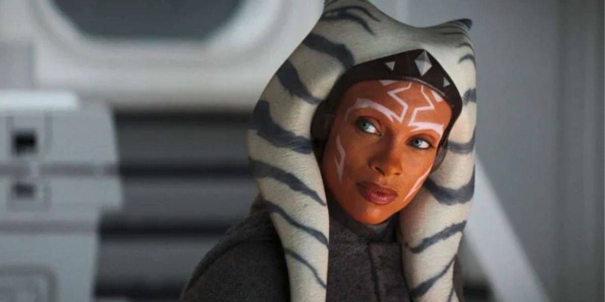 A person with orange skin, white facial markings, and a headpiece with long, striped lekku looks attentively to the side. They are wearing a dark robe, set against a gray, futuristic background.