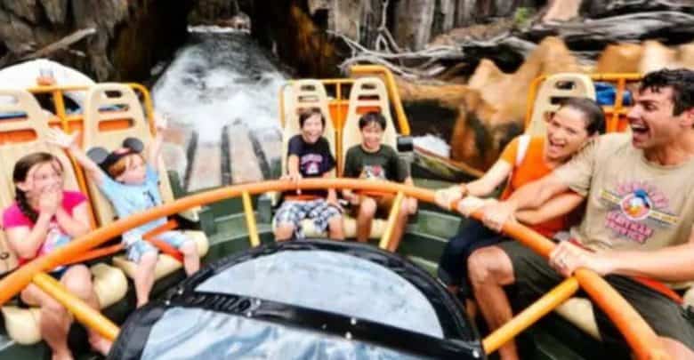 A group of six people, including a lively cast member, are on a water ride, holding onto a circular raft. They appear excited and are laughing as water splashes around them. The ride is surrounded by rocky terrain, and they are approaching a tunnel.