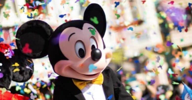 A person in a Mickey Mouse costume stands surrounded by colorful confetti at Disneyland. The character is wearing a black jacket, white shirt, and yellow bow tie. The background is blurred, creating a festive and cheerful atmosphere.