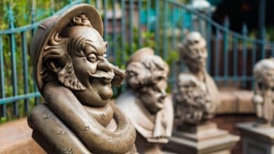 A close-up of whimsical stone busts with exaggerated facial features, displayed outdoors in a row—each one seeming to express its own guest pet peeves—framed by a decorative iron fence and lush greenery in the background.